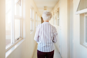 Senior, back and woman walking with corridor in retirement home by window for morning in apartment. Elderly female person or resident having a stroll down hallway, passage or path in flat or building