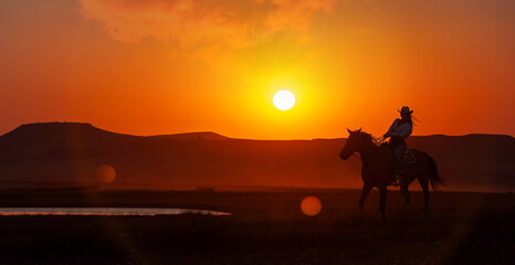 Fototapeta premium silhouette cowgirl in the sunset riding in the prairie near a waterhole