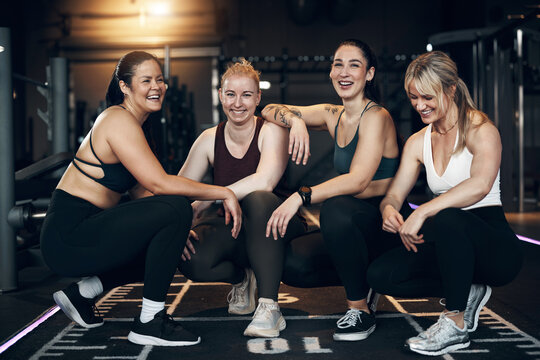 Laughing group of diverse young women in sportswear squatting on the floor of a gym after an exercise class together