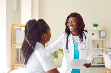 Fototapeta premium Portrait of smiling friendly female african american doctor therapist talking with a young woman patient holding report file with appointment standing in office. Medicine and health care concept.