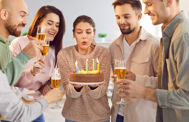 Cheerful young happy woman blowing out the candles on her birthday cake standing in the living room at home at evening party with her best friends holding glasses of champagne. Birthday party concept