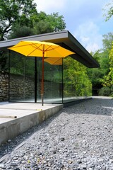 A minimalistic pavilion made of grey steel and glass, set against a grey gravel path, showcases a bright yellow umbrella propped open