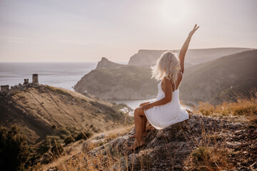 Happy woman is sitting on a hillside, wearing a white dress. She is surrounded by a beautiful landscape, with a body of sea in the background. Concept of peace and happiness.