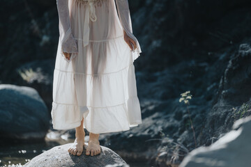 Woman in white dress standing barefoot on a rock in natural setting, evoking serenity and...