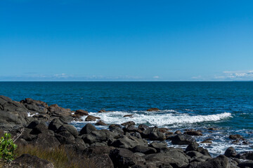 Rocky beach on a bright sunny day. Rocky shoreline with turquoise water and cloudless sky. Many Bay, Raglan, New Zealand
