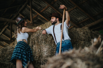 Young couple dressed in casual clothes and cowboy hats, leaning on hay bales in a rustic barn, enjoying a moment in a relaxed, rural setting. © qunica.com