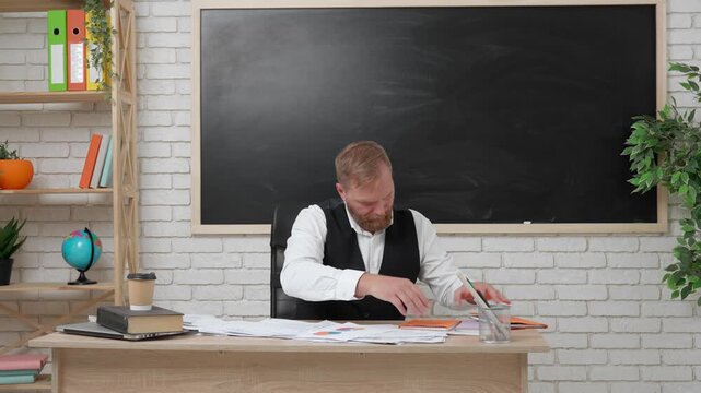 Man college teacher sit at desk in classroom, searching for paper in the pile of sheets and books on the table, tired expression. Education concept.