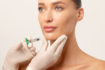 Beautiful European lady getting facial injection in chin, cosmetologist holding syringe, white...