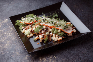 Summer salad with pieces of bacon, fresh cucumbers, cheese cubes and mushrooms. Green salad on a black plate on a dark background. Free space for an inscription. View from above.