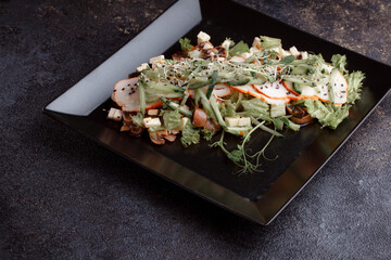 Summer salad with pieces of bacon, fresh cucumbers, cheese cubes and mushrooms. Green salad on a black plate on a dark background. Free space for an inscription. View from above.