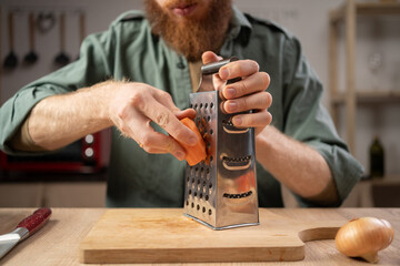 Bearded man rubs carrots on a grater in his home kitchen, concept of healthy eating
