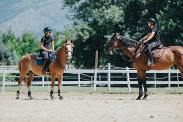 Two horse riders in a sunny outdoor arena, both wearing helmets and engaging in a friendly...