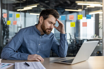 Tired and upset young man sitting in the office at the desk in front of the laptop, looking worriedly at the screen and holding his head with his hand