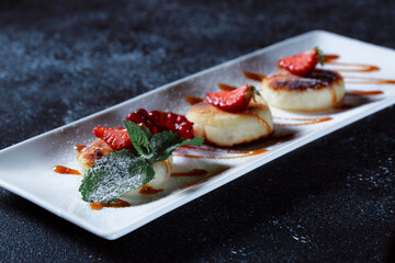 Cottage cheese pancakes decorated with strawberries and red currants. Beautiful serving of the dish on a textured dark blue background. Restaurant dessert menu.