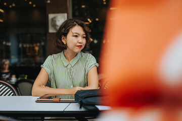 An Asian woman in a green striped shirt sits thoughtfully at a cafe table, with a tablet and bag in front of her.