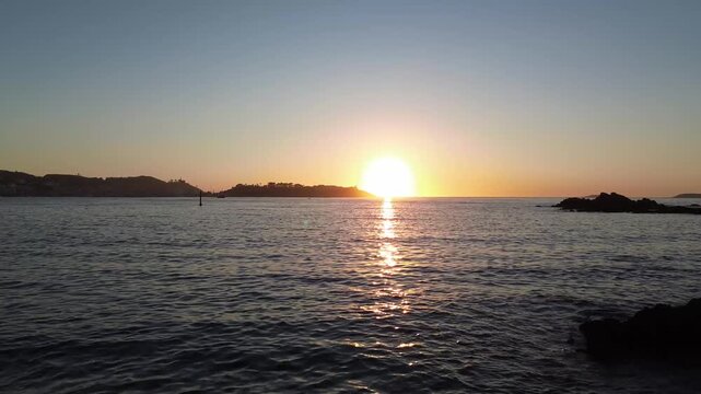 Camera glides from Monte Lourido over calm waters at sunset, capturing the silhouette of the Parador de Baiona against the orange sky. The sun sets beside Baiona as it reflects on the tranquil sea.
