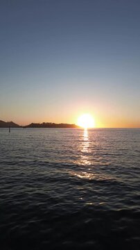 Camera glides away over the calm waters towards Monte Lourido, capturing the silhouette of the Parador de Baiona against the orange sky. The sun sets beside Baiona as it reflects on the tranquil sea.