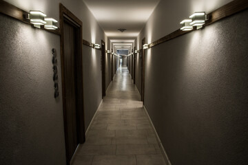 A long, narrow corridor with multiple doors on both sides, illuminated by wall-mounted lights, creating a sense of depth and perspective.
