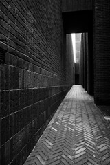 A narrow alleyway between two brick buildings, leading to a brighter area, with a strong contrast between the dark alley and the light at its end.