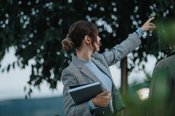 A businesswoman in a plaid blazer points while holding a notebook during an outdoor meeting,...