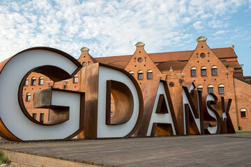A striking outdoor installation featuring the name GDANSK in oversized 3D letters set against the historic architecture of the city under a clear blue sky.