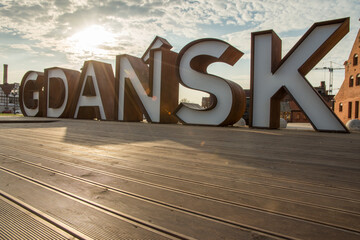 A striking outdoor installation featuring the name GDANSK in oversized 3D letters set against the historic architecture of the city under a clear blue sky.