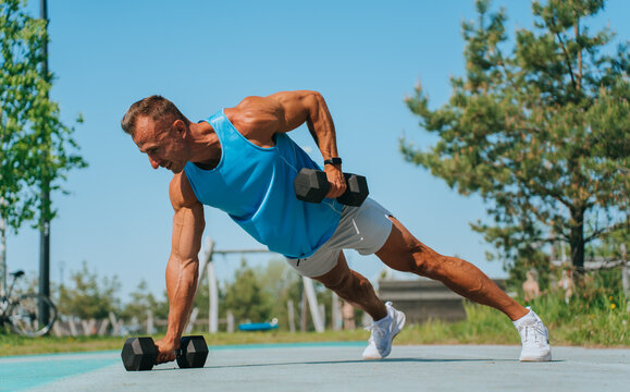 man performs a renegade row exercise with dumbbells outdoors, showcasing strength and focus in a park setting