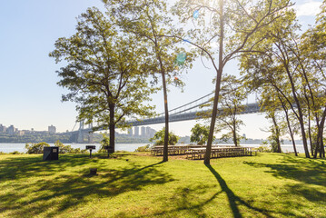 Picninc tables and barbeque grills on grass among trees in a riverbank park on a sunny autumn day. A lage supension road bridge is in background.