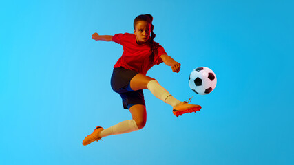 Female soccer player mid-flight, striking ball with precision and power in neon light against colorful gradient background. Concept of women and sport, team games, movement, victory, action. Ad