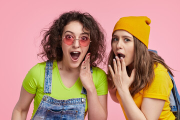 Two young women in casual outfits, expressing surprise while gossiping. They are posing against a pink background, showcasing a fun and lively moment.