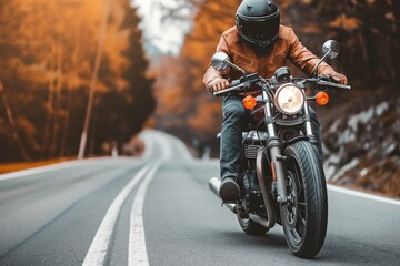 Naklejka premium A motorcyclist in a leather jacket rides down a winding road with autumn foliage and a forest backdrop.