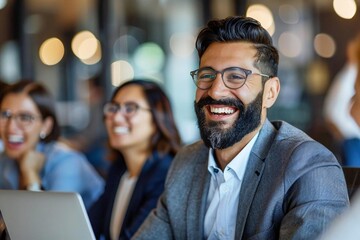 Fototapeta premium Young businessman with a well-groomed beard video conferencing with colleague on laptop, both laughing, while some people wear eyeglasses in office.