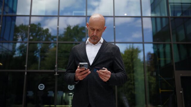 Bald businessman dressed in dark jacket is standing in front of modern glass office building. The man is holding smartphone in one hand and a credit card in other, trying to make online purchase.