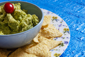 Guacamole in a blue bowl with a cherry tomato surrounded by nachos on a plate decorated with flowers