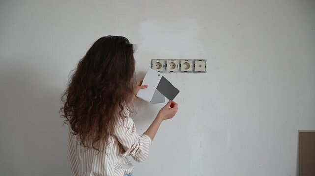 Woman comparing paint color swatches against a wall with electrical outlets. Home renovation project, choosing color scheme for interior design