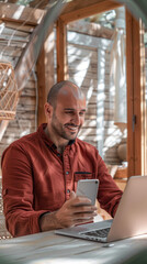 Fototapeta premium A man in a red shirt is smiling while using his cell phone and laptop