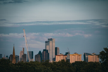 Fototapeta premium Skyline of a modern city featuring high-rise buildings, construction cranes, and architectural landmarks, framed by trees under a serene, cloud-streaked sky.