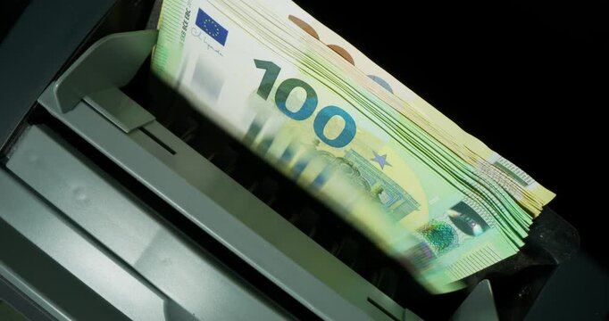 A man's hand puts euro bills of different denominations into a calculating machine. The machine counts banknotes standing on a black background. Close-up