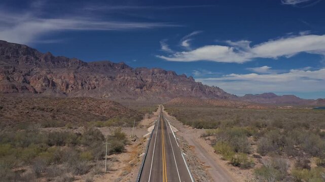 Aerial view of a desert highway leading to a mountain range, showcasing stunning scenery with dynamic cloud formations under a bright blue sky. Road to Loretto city in Mexico