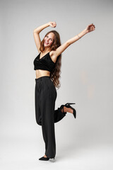 Young Woman in Black Crop Top and Pants Posing With Arms Raised Against a White Wall