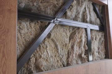 Close-up of insulation material fitted between metal frames within a wooden structure. The texture of the insulation and the metal supports contrasts against the natural wood finish of the frame