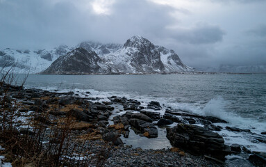 A scenic coastal view featuring a snow-capped mountain, rocky beach, stormy sky, and dynamic ocean, Lofoten, Norway