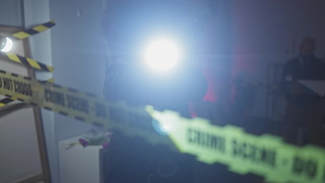 Silhouetted detectives with flashlights investigate a crime scene within a blurry living room interior, marked by caution tape.