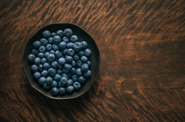 Ripe blueberries in a dark plate on a wooden background with copy space. Fresh blue berries in a bowl. Top view, flat lay.