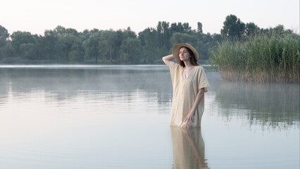 A dreamy girl or young woman in a hat and a simple dress stands in a lake amidst the mist, holding her hand behind her head