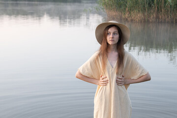 A dreamy girl or young woman in a hat and a simple dress against the backdrop of a lake and mist