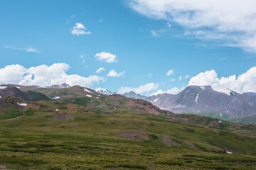 Dramatic alpine view from green hills to large rocky mountain range with glacier under clouds in blue sky. Scenic landscape with big snow mountain top. Changeable cloudy weather in high mountains.