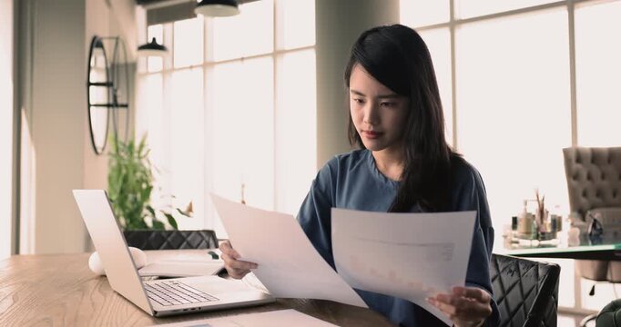 Workflow of focused Asian woman office employee, accountant, secretary or female lawyer at workplace, sit at desk with laptop hold papers, learn legal document, review terms, conditions, do paperwork