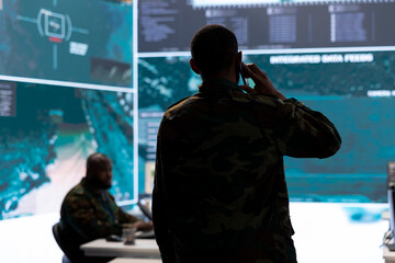 Military man answers phone call in a mission control center, working with advanced technology and satellite imagery to track potential threats and monitor real time data. Intelligence activities.