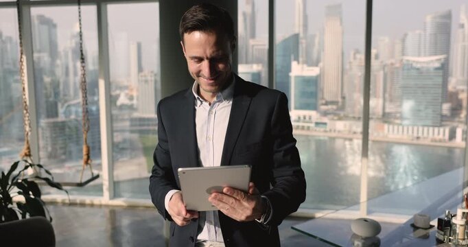 Optimistic successful businessman, company owner using digital tablet working chatting on-line, answers on email, solve business remotely in contemporary office with city view from panoramic window